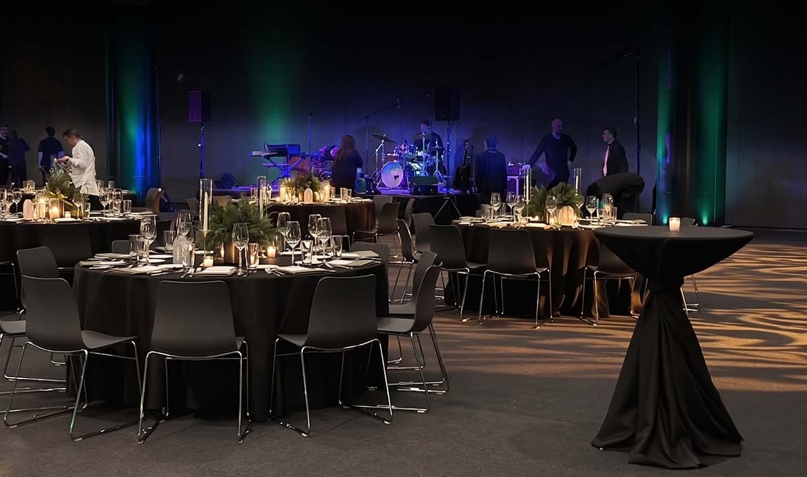 a stage with a band setting up in the background and banquet tables in the foreground. Black curtains behind the stage and black table cloths on the tables.